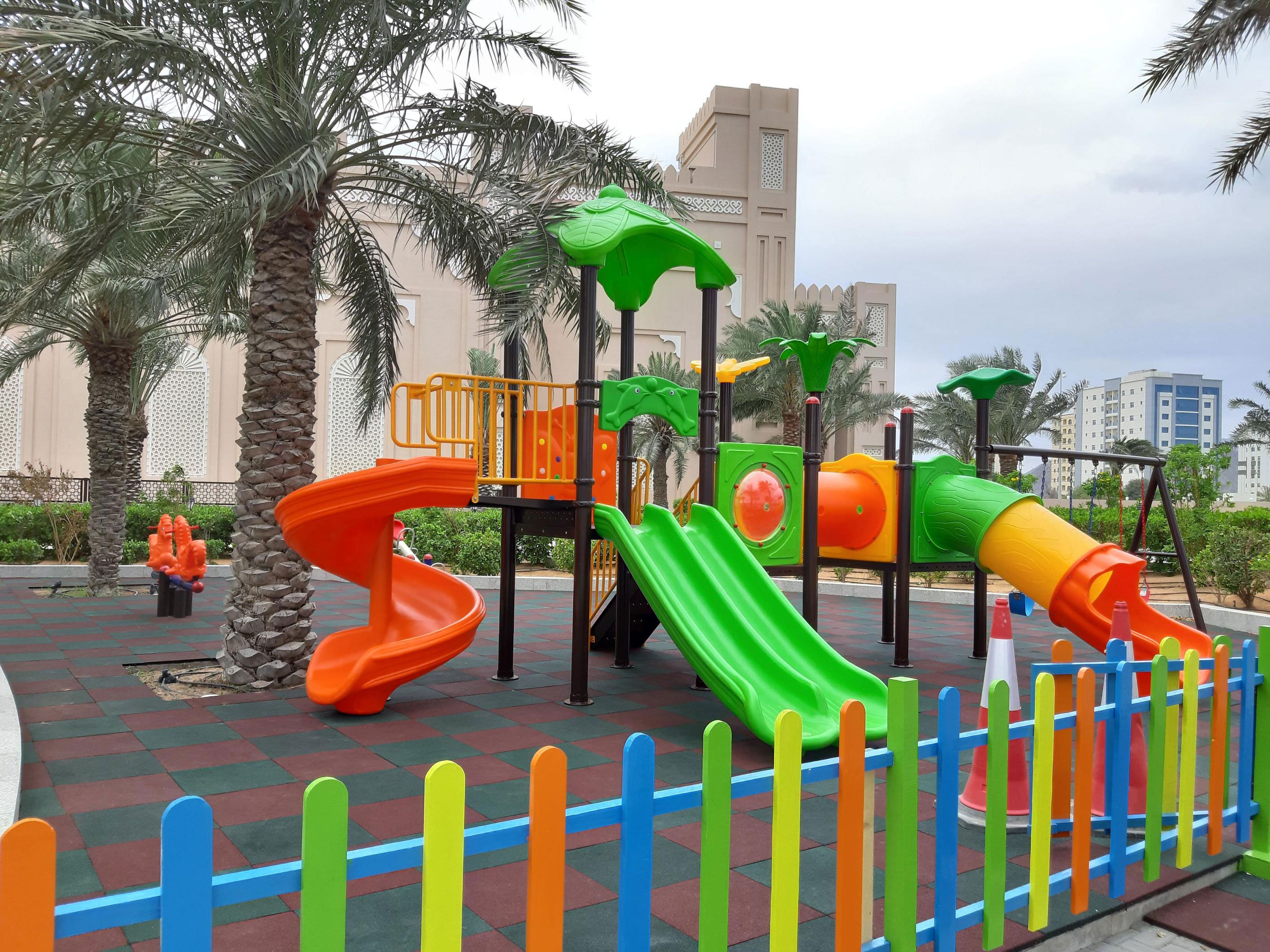 Un parque infantil con toboganes y estructuras para escalar, rodeado de palmeras y una zona pavimentada.