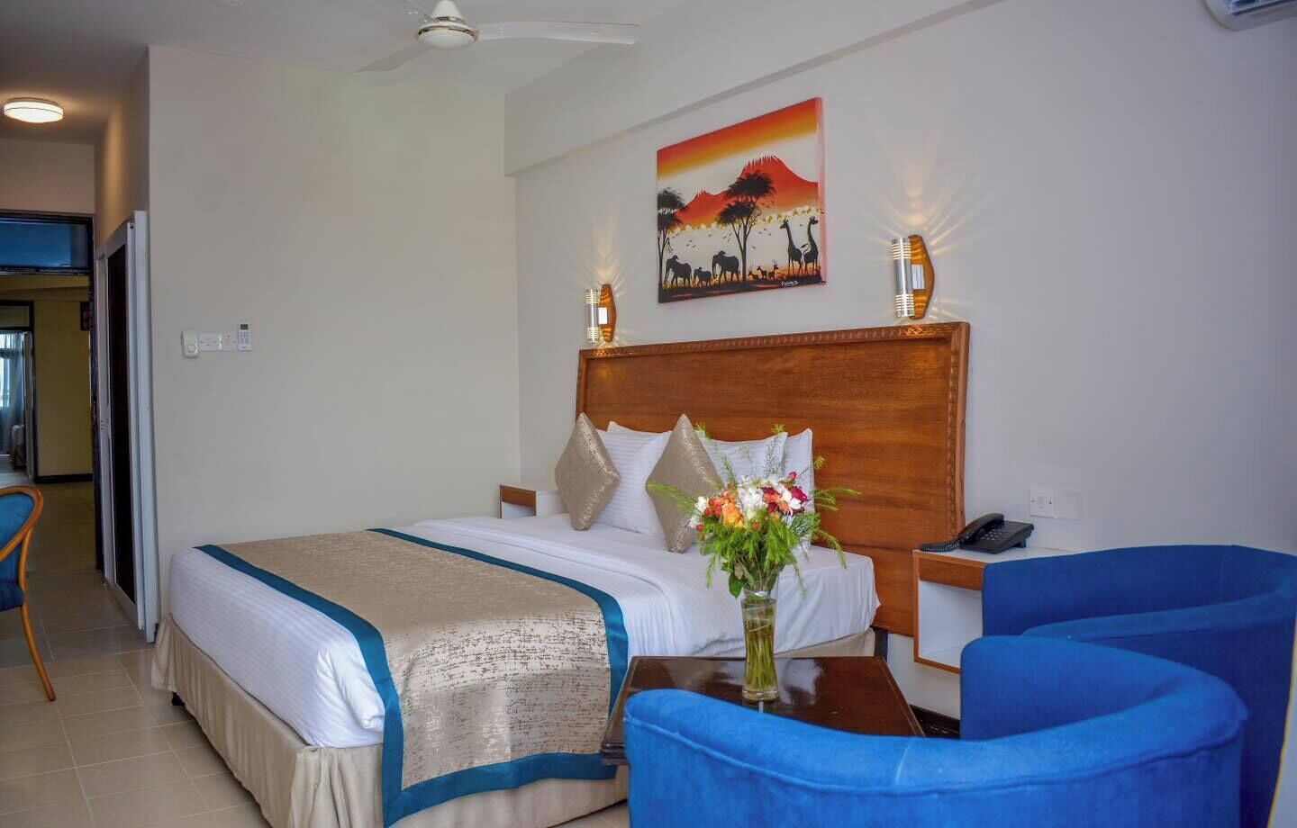 A hotel room with a bed, a desk with a phone, two chairs, a painting on the wall, and a ceiling fan.