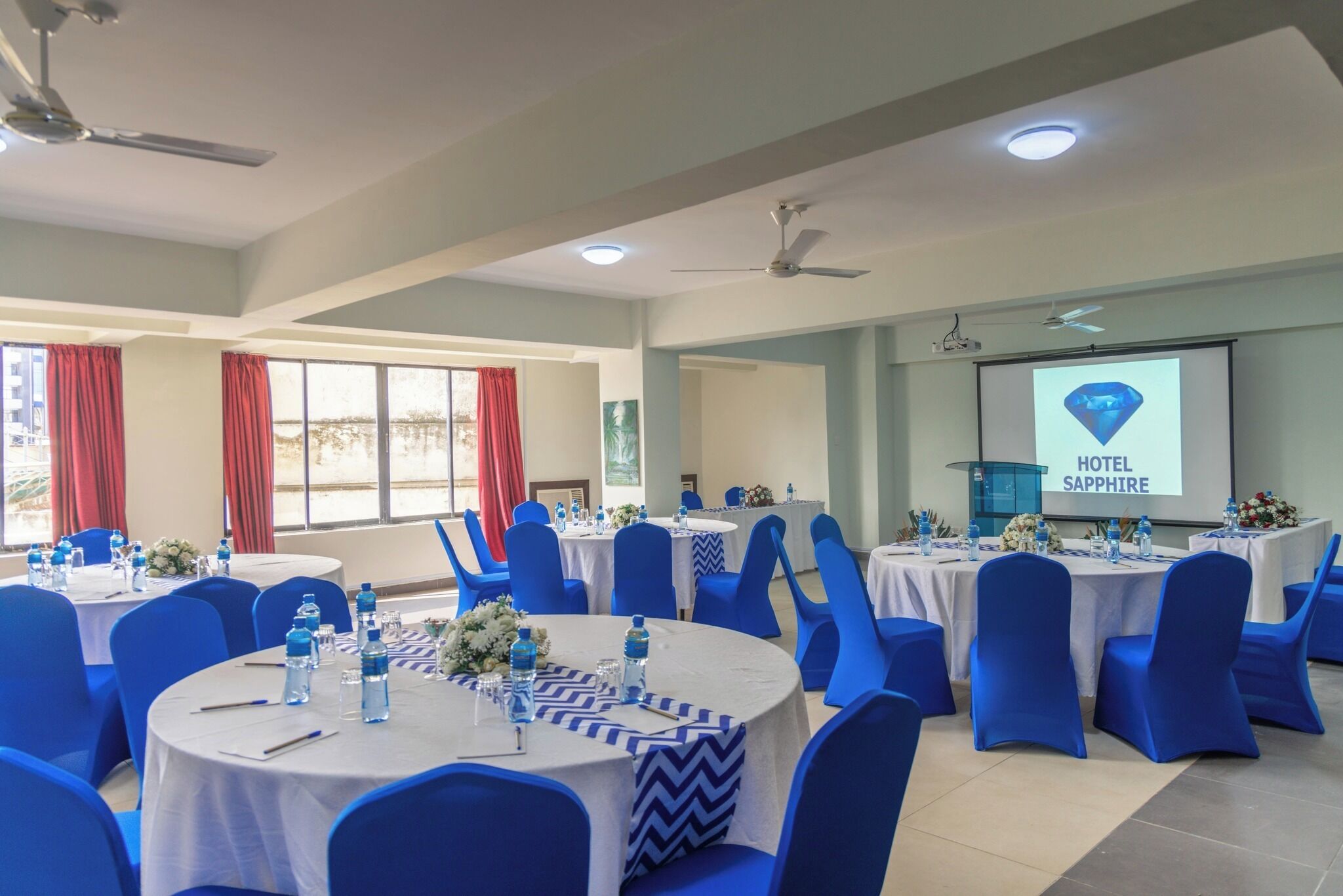 A conference room with round tables, blue chairs, and a projector screen displaying 'HOTEL SAPPHIRE'.