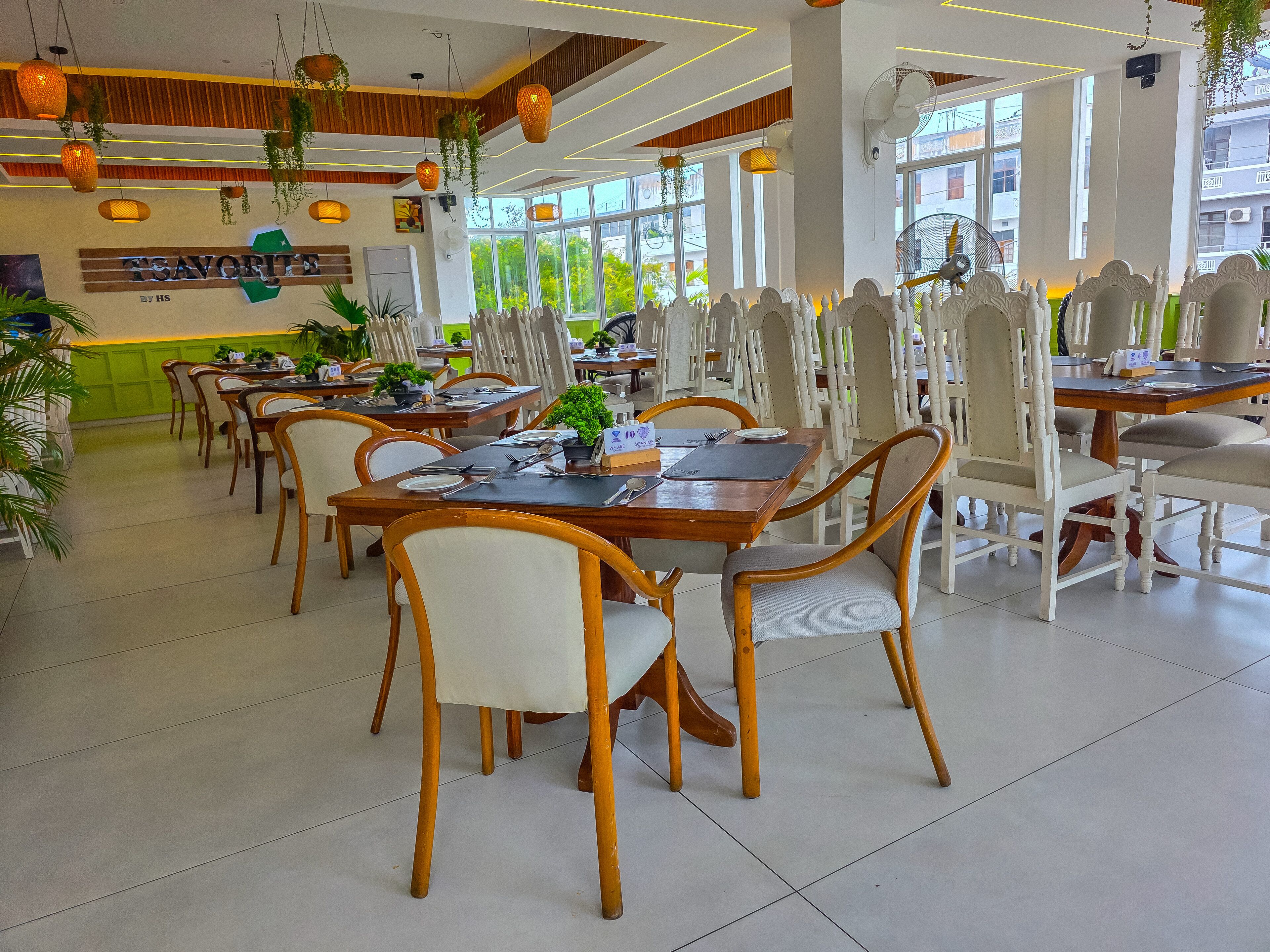 Un restaurante con mesas y sillas de madera, plantas colgantes y amplios ventanales.