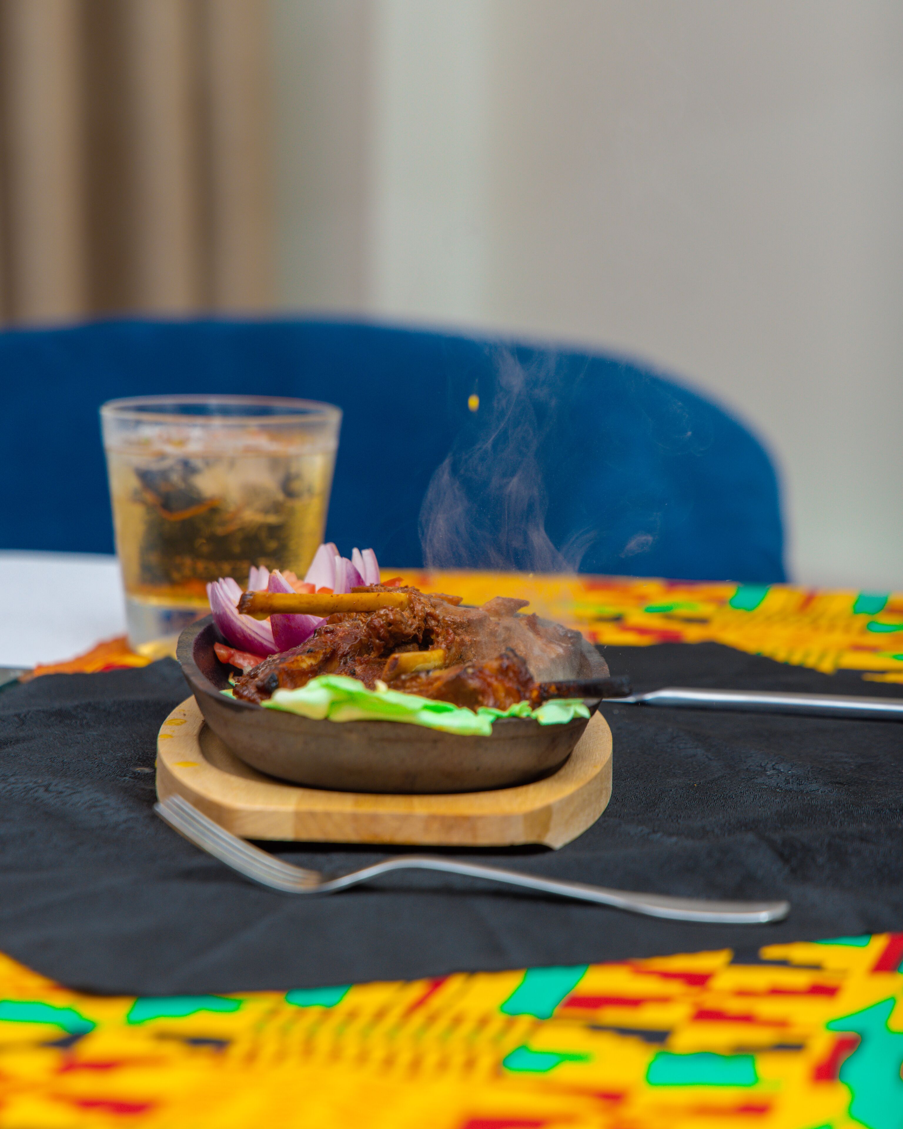 A meal with a bowl on a wooden board, a glass of beverage, and a colorful tablecloth.