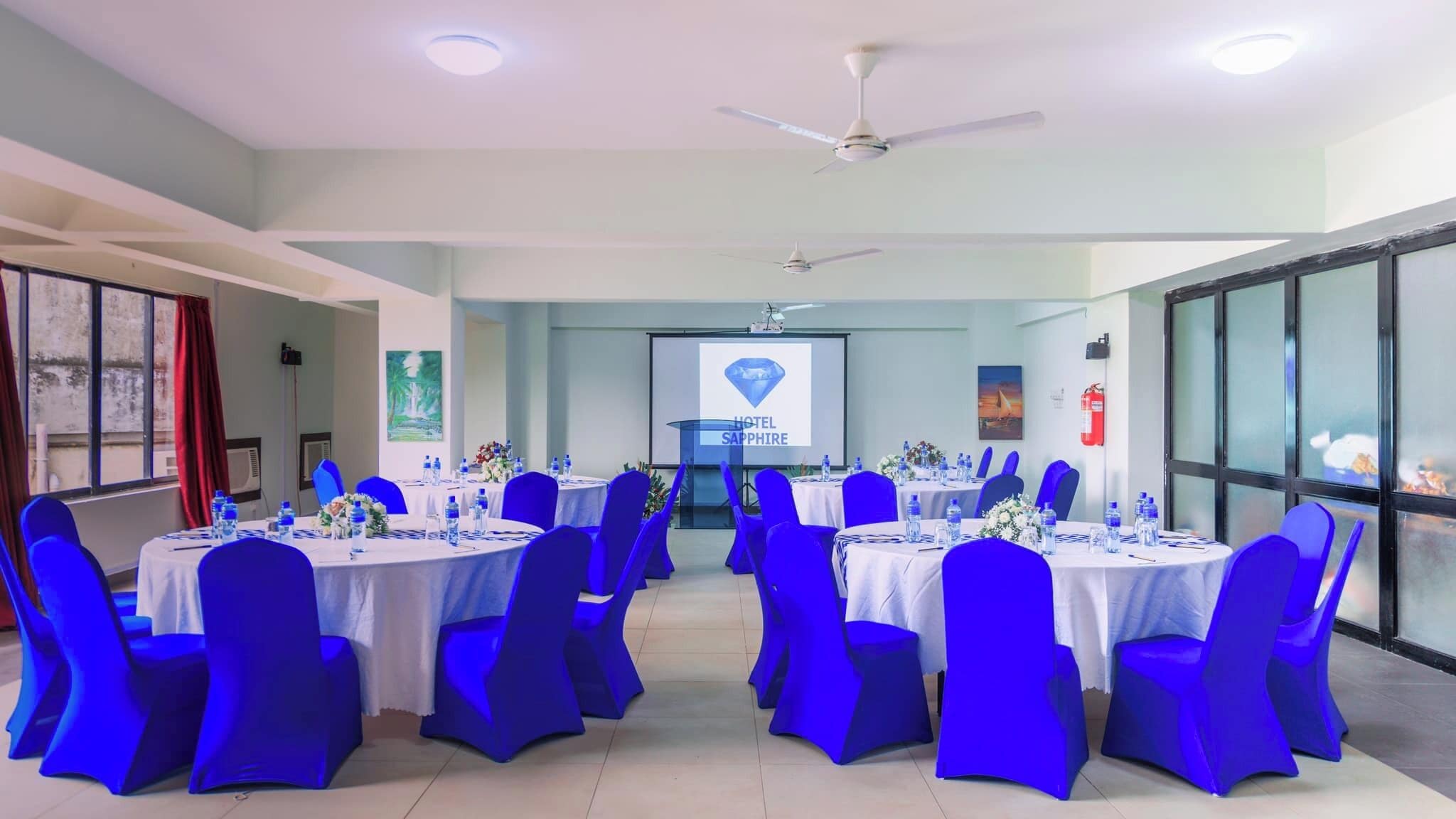 A conference room with round tables, blue chairs, and a projector screen displaying 'HOTEL SAPPHIRE'.