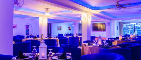 A restaurant with blue chairs and tables, a ceiling fan, and a TV screen.