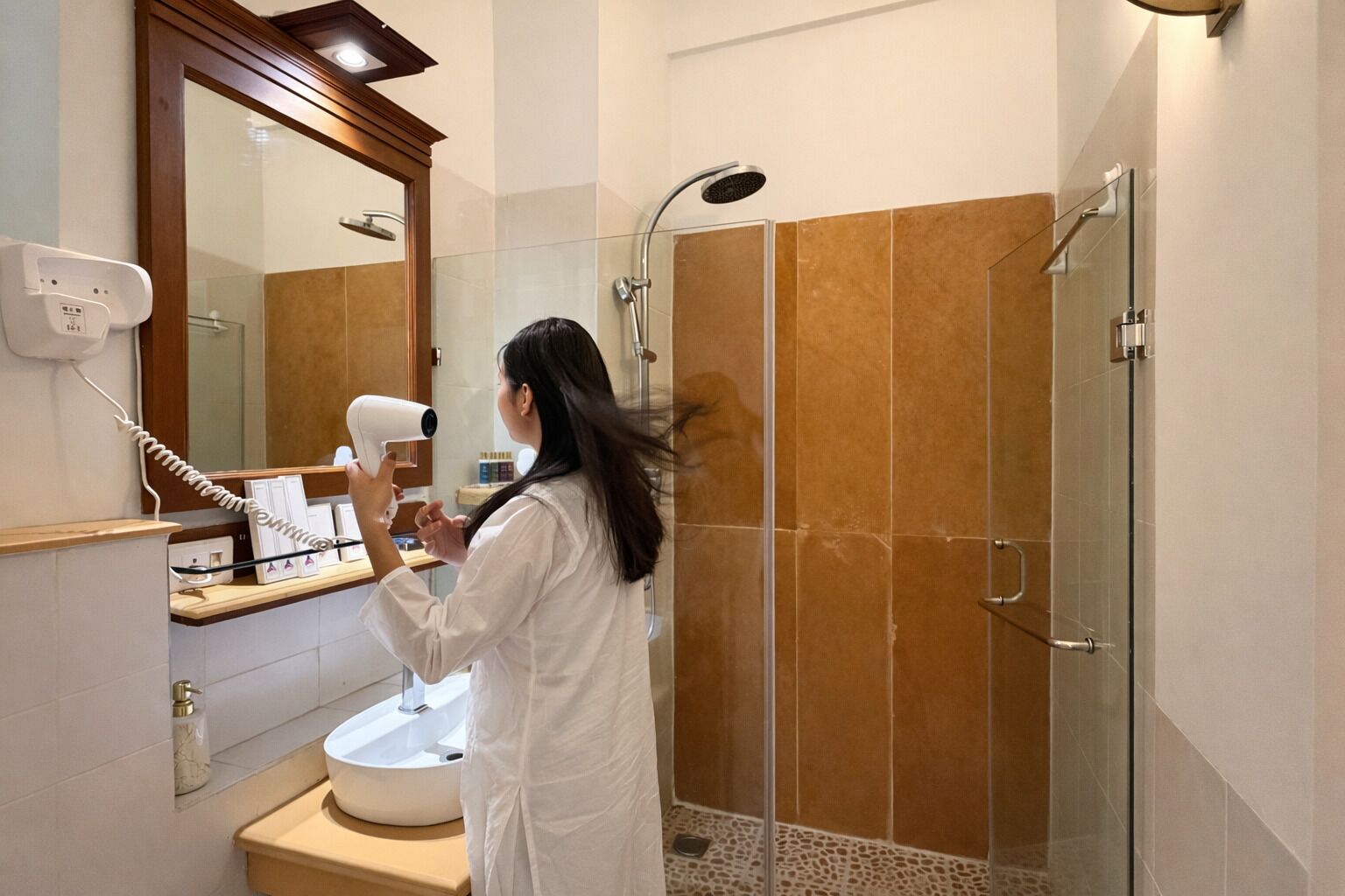 A person in a white robe using a hairdryer in a bathroom with a glass shower enclosure and a wooden-framed mirror.