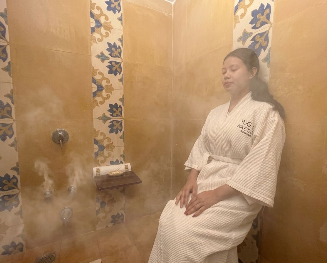 A person in a robe sitting in a steam room with patterned walls and a towel rack.