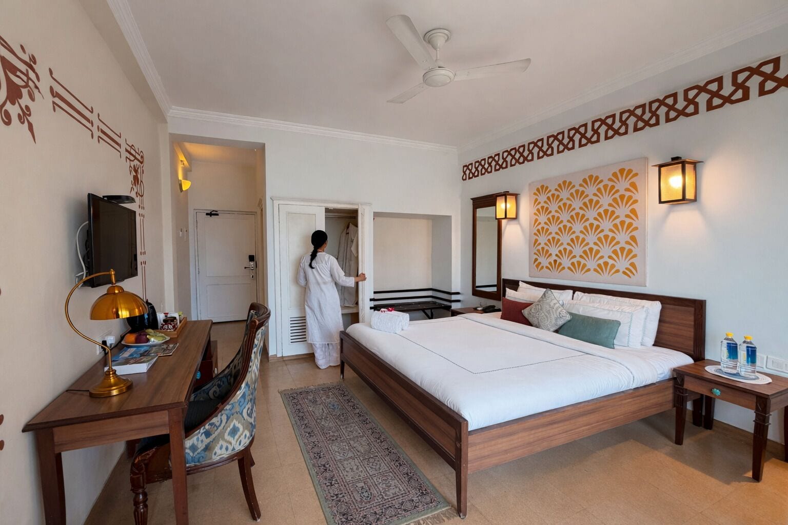 A hotel room with a bed, bedside tables, a desk with a lamp, a chair, a TV, a wardrobe, and a patterned wall design.