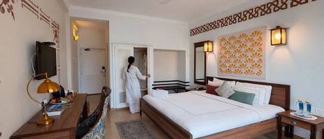 A hotel room with a bed, bedside tables, a desk with a lamp, a chair, a TV, a wardrobe, and a patterned wall design.