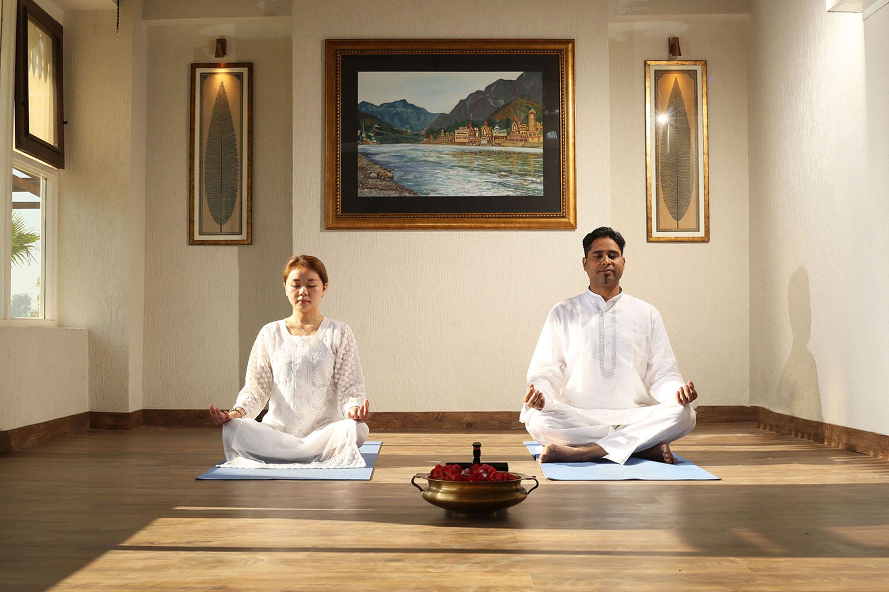 Duas pessoas meditando em tapetes azuis em um quarto com piso de madeira, obras de arte emolduradas nas paredes e uma tigela de latão com flores no chão.