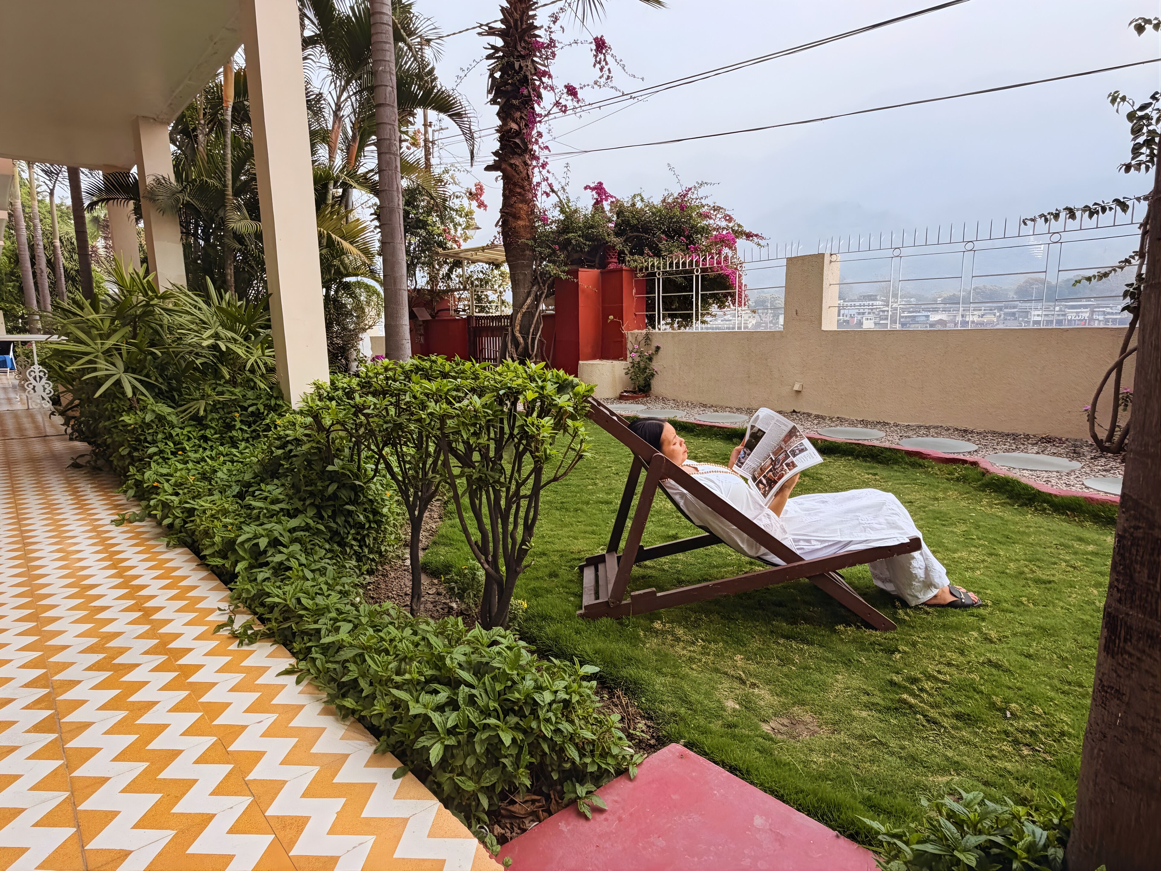 A covered outdoor area with a tiled walkway, a grassy lawn, and a lounge chair with a magazine.