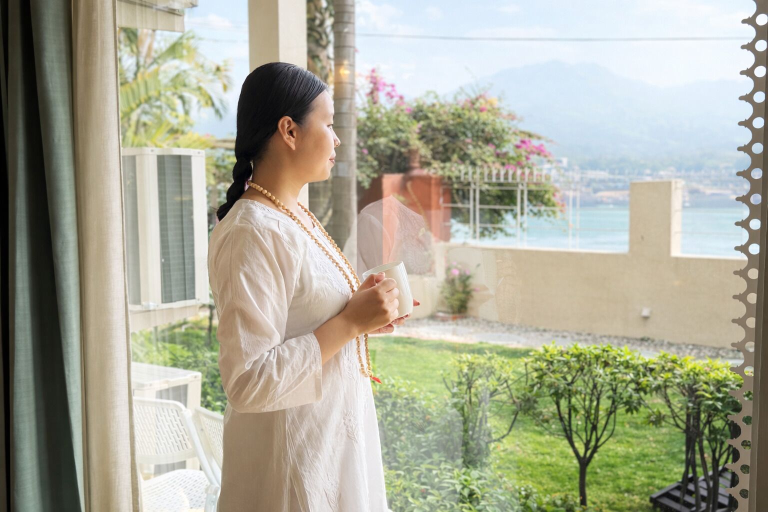 Una persona de pie junto a una ventana con vista a un jardín y un cuerpo de agua.