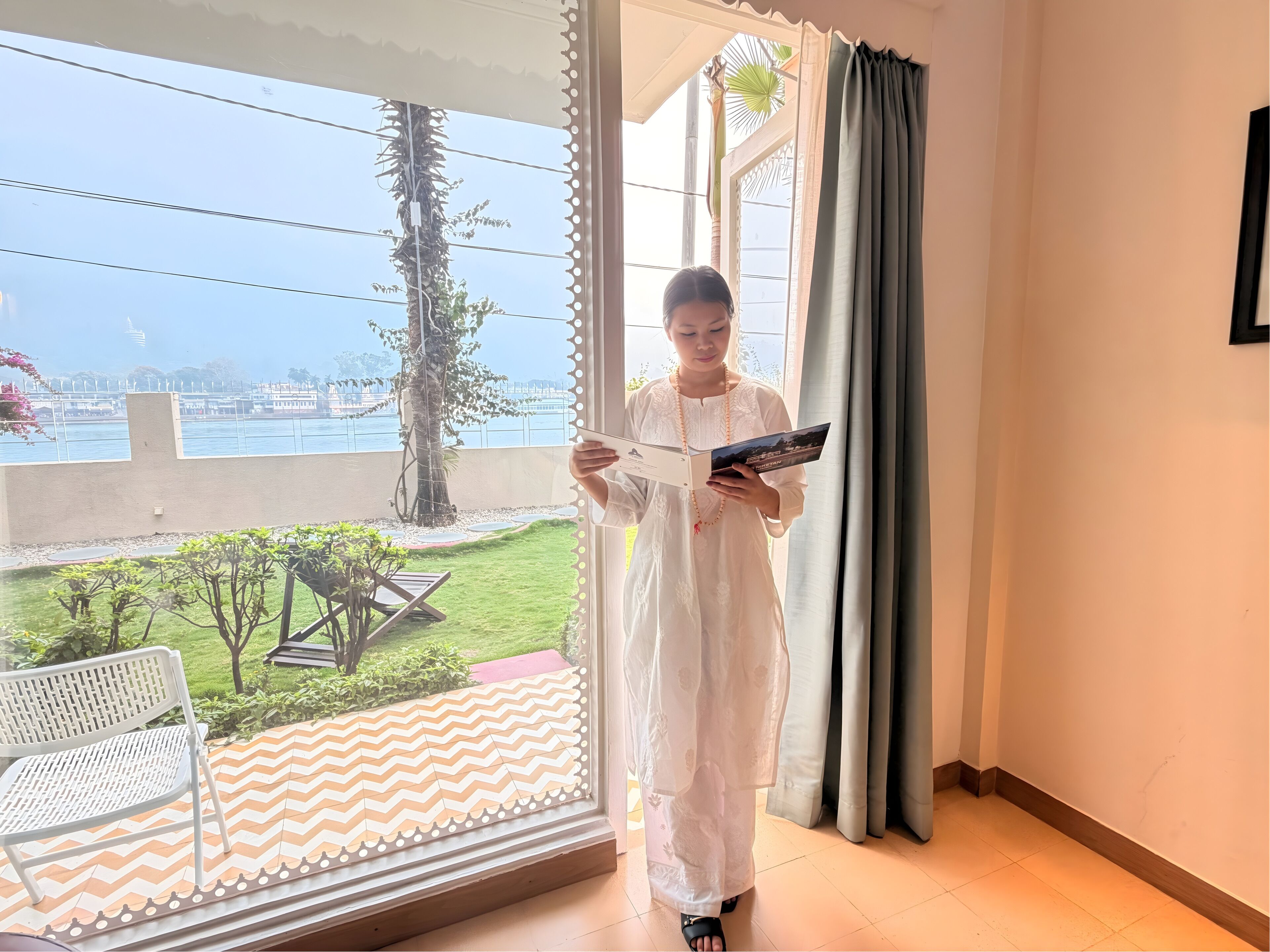 A person standing by a window with a view of a body of water and greenery, holding a booklet.
