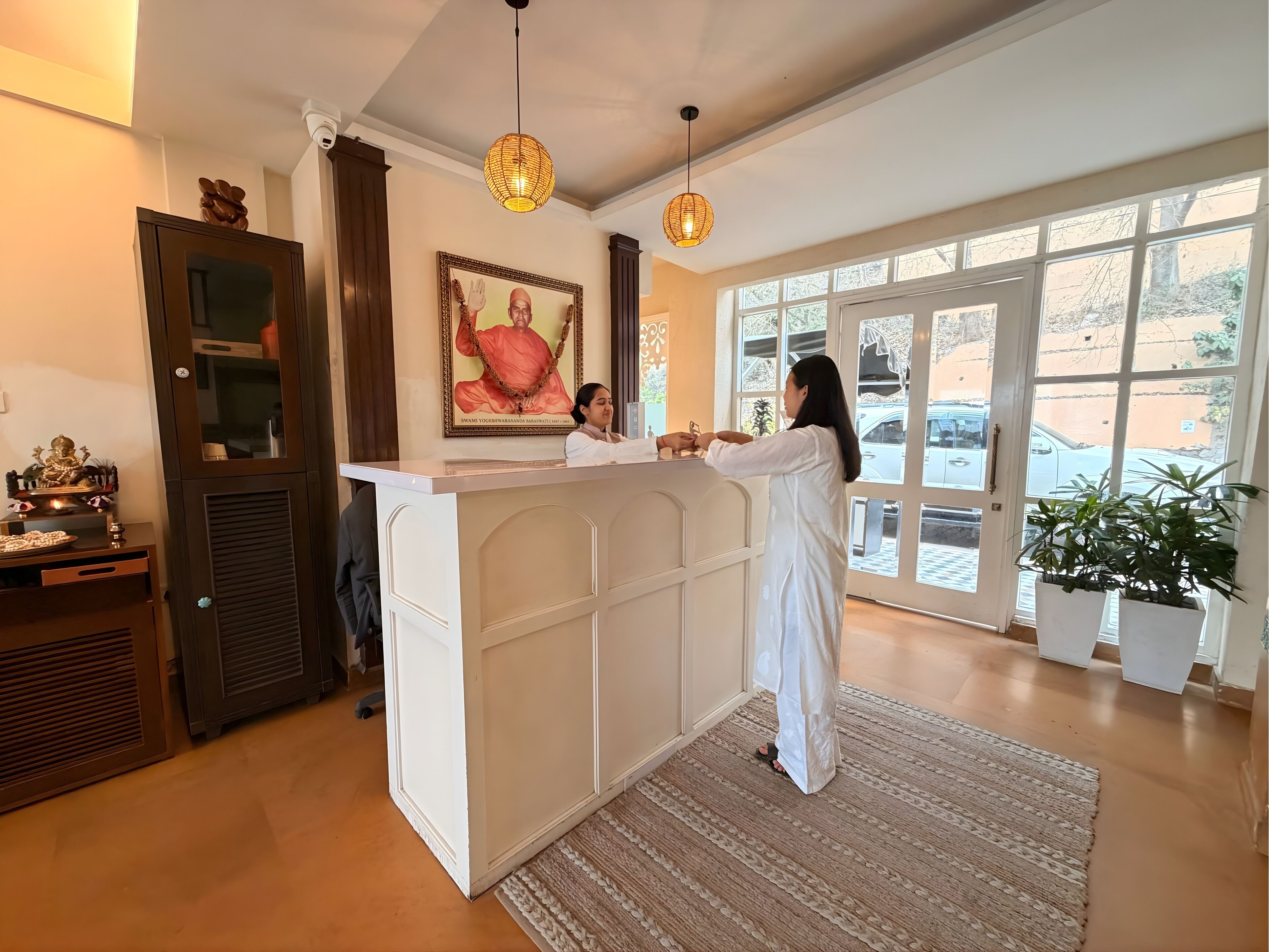A reception desk with a framed picture above it, a large window with a view of the outside, and a potted plant to the right.