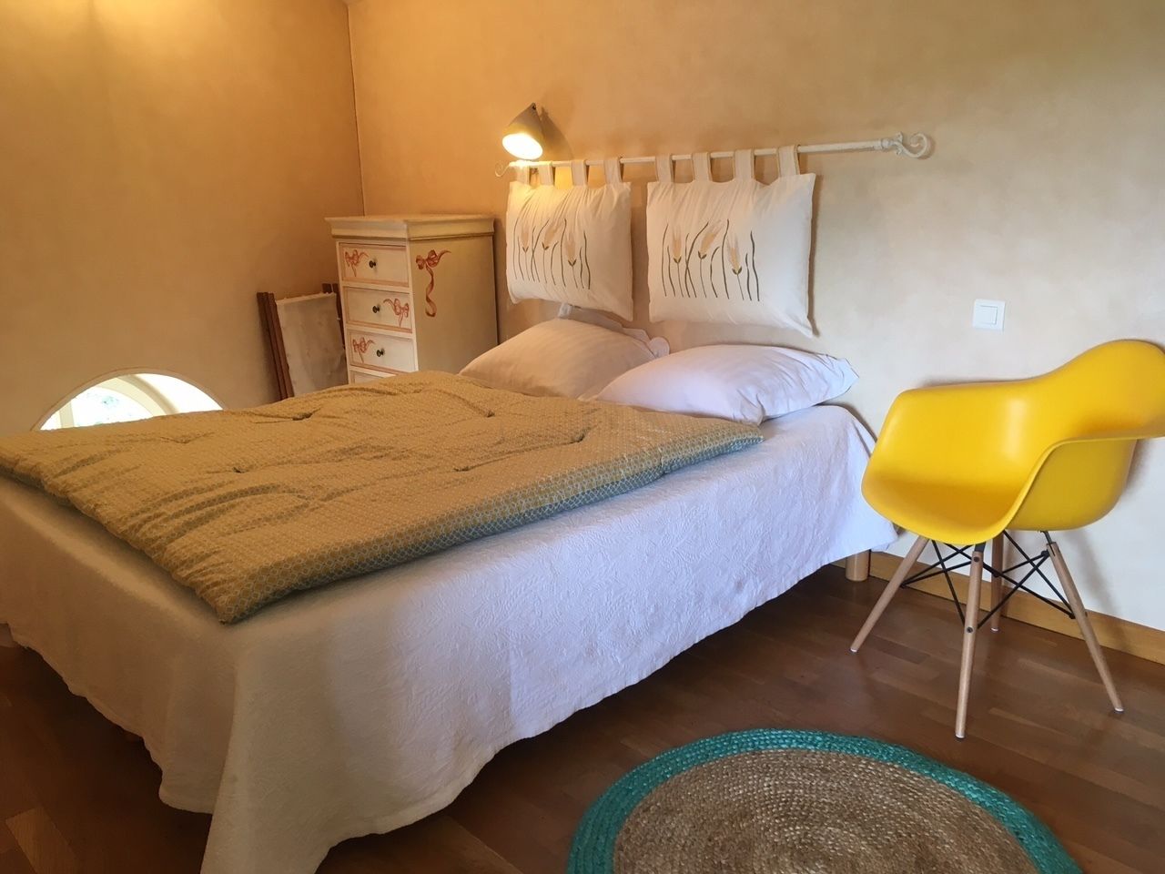Een slaapkamer met een bed, een gele stoel, een nachtkastje en een wandlamp.