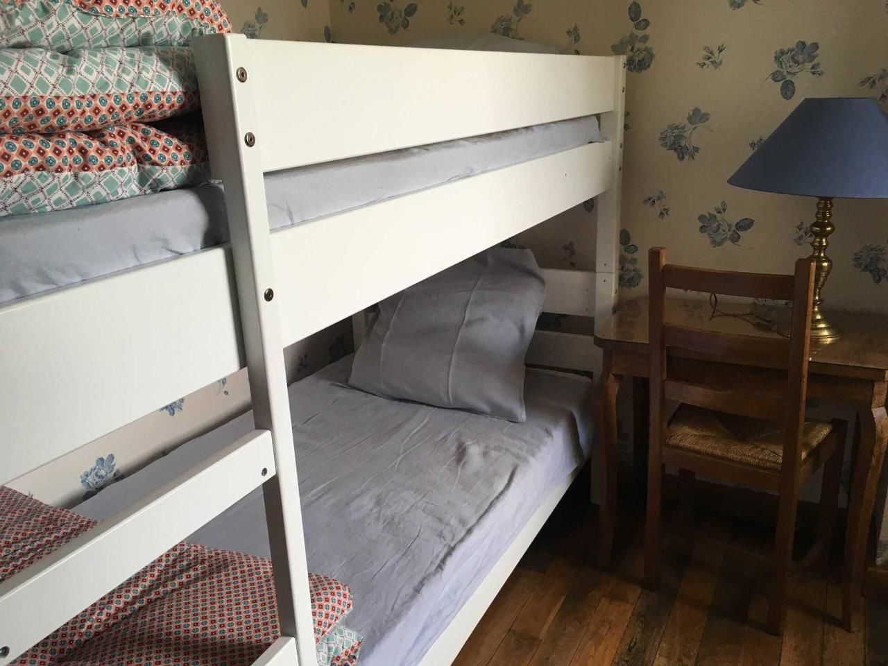 Ein Etagenbett mit grauer Bettwäsche, ein Holztisch mit einer Lampe und ein Stuhl.