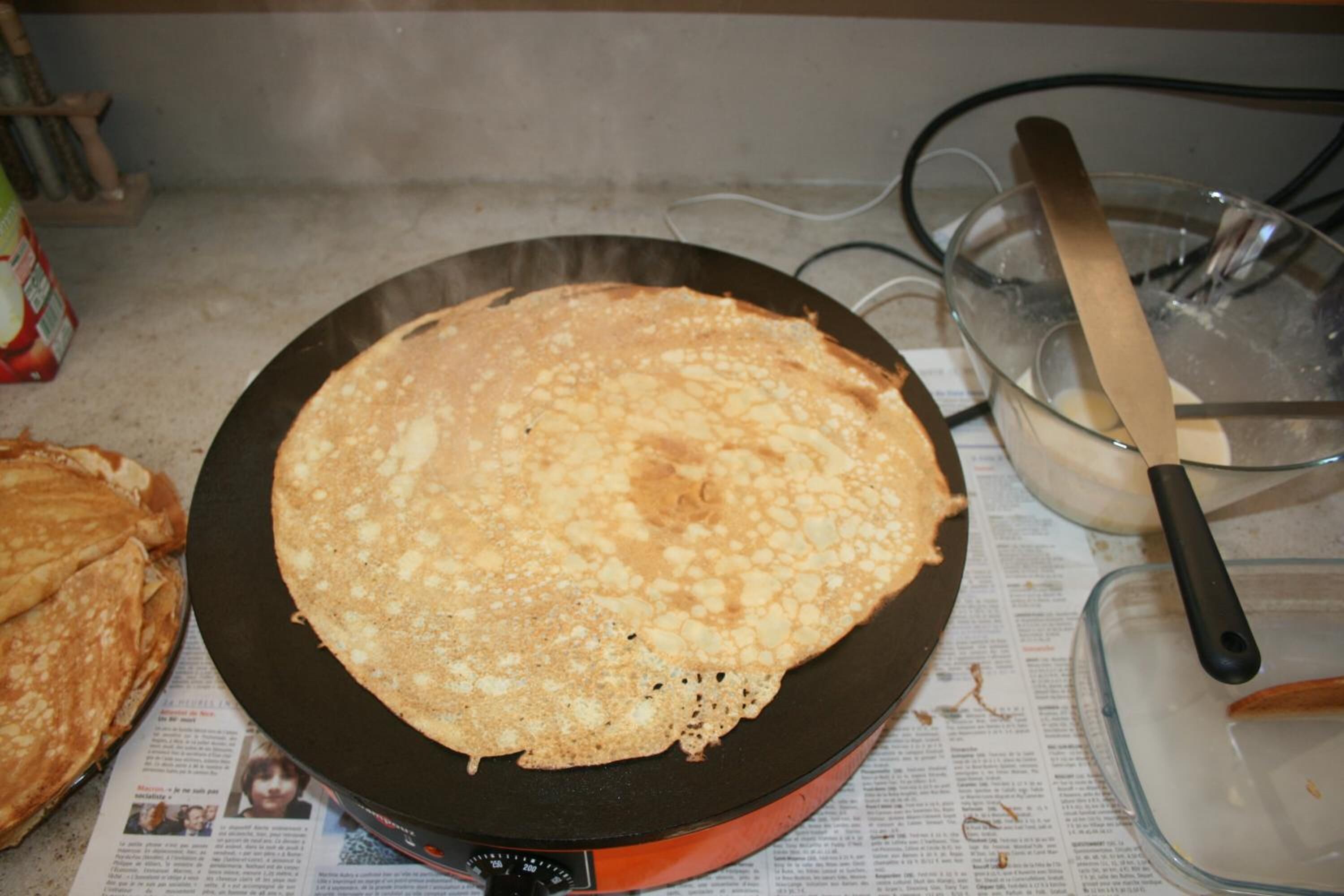A crepe on a pan, a knife, and a glass bowl on a kitchen counter.