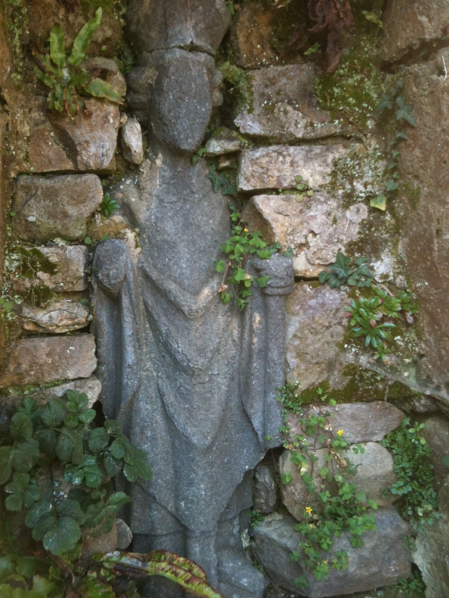 A stone statue of a figure with outstretched arms, set against a stone wall with moss and small plants growing around it.