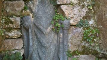 A stone statue of a figure with outstretched arms, set against a stone wall with moss and small plants growing around it.