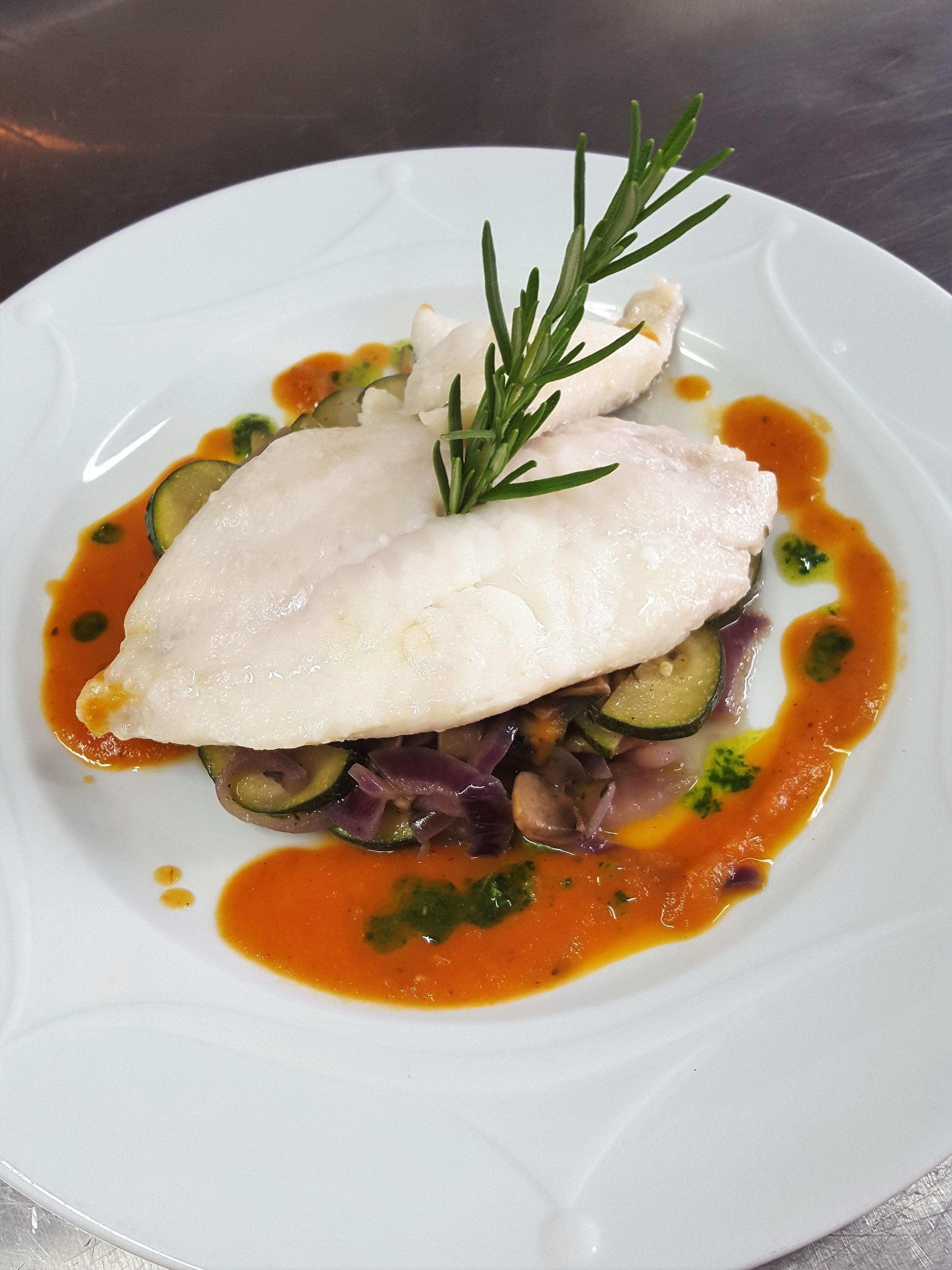 A white plate with a piece of white fish, zucchini, red onion, and a sprig of rosemary.
