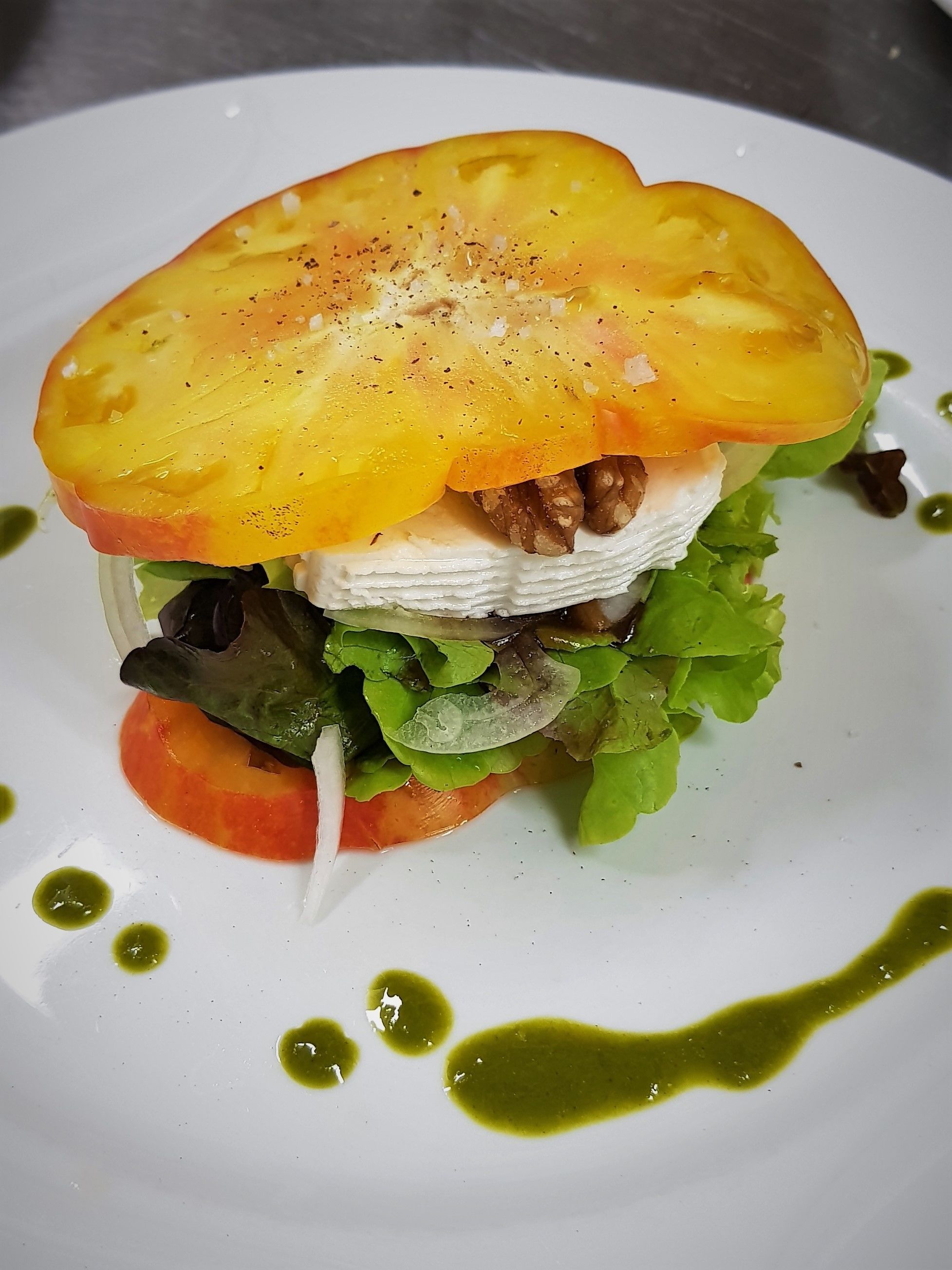 A sandwich with a slice of tomato on top, lettuce, and a creamy filling, served on a white plate with green sauce.