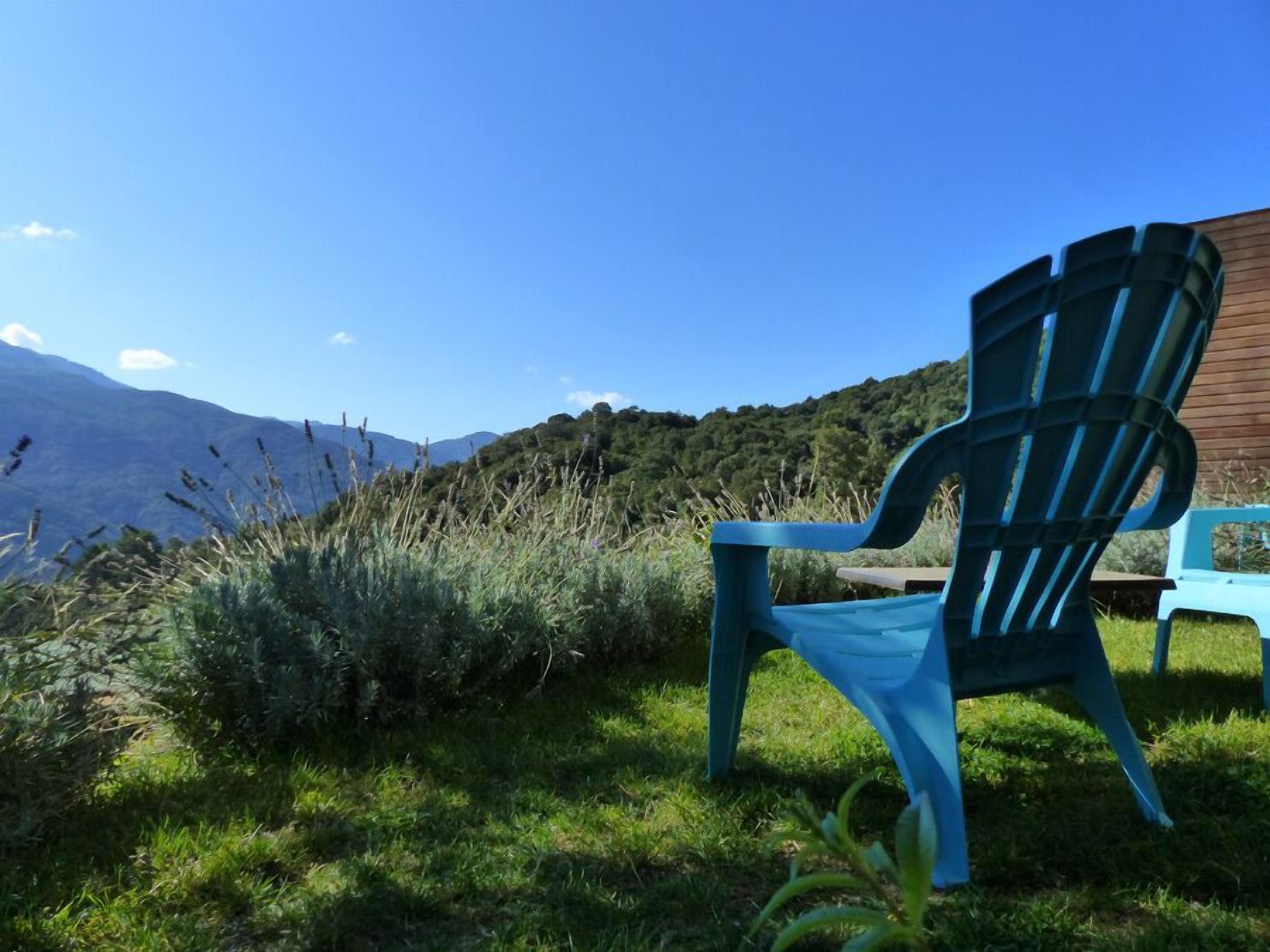 Une chaise bleue, placée à l’extérieur, dans un espace verdoyant, avec une vue sur des montagnes et des collines en arrière-plan.