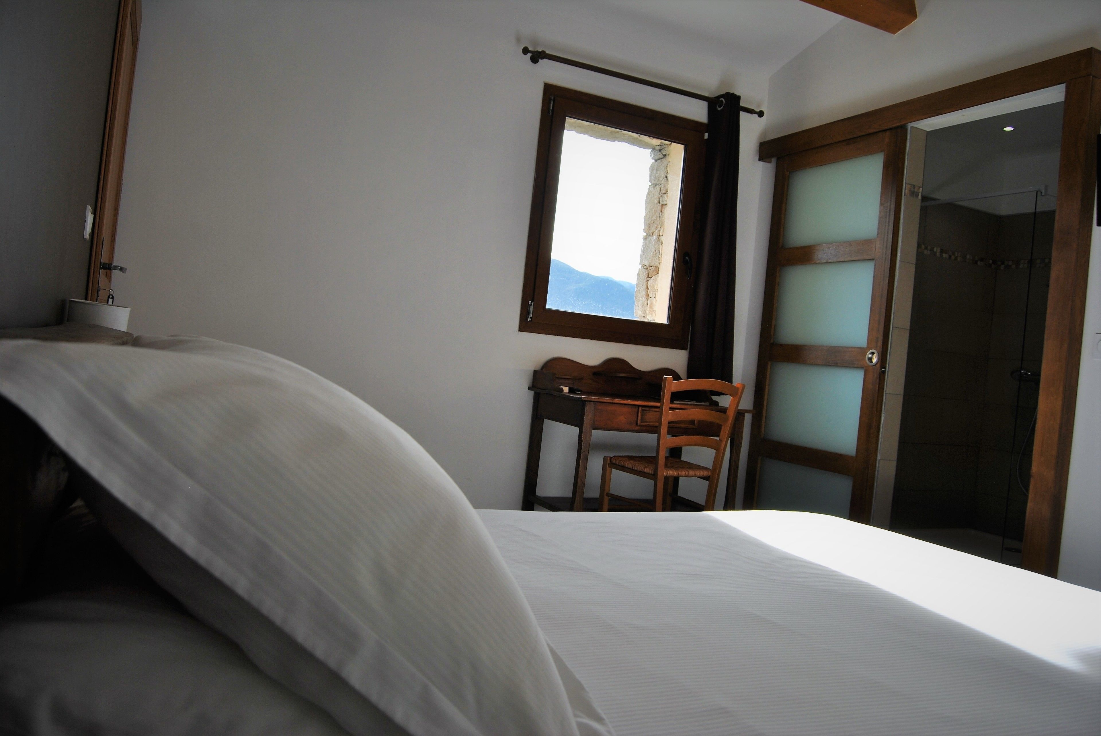 A bedroom with a bed, a desk, and a chair. There is a window with a view of the sky and a door with frosted glass panels.