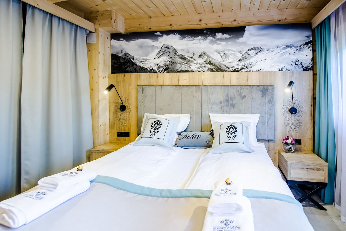 Cama doble con sábanas blancas, dos almohadas con diseños florales y una almohada que dice 'Relax'. Tiene cabecera de madera, mesita de noche con lámpara y un mural en la pared con paisaje de montaña.