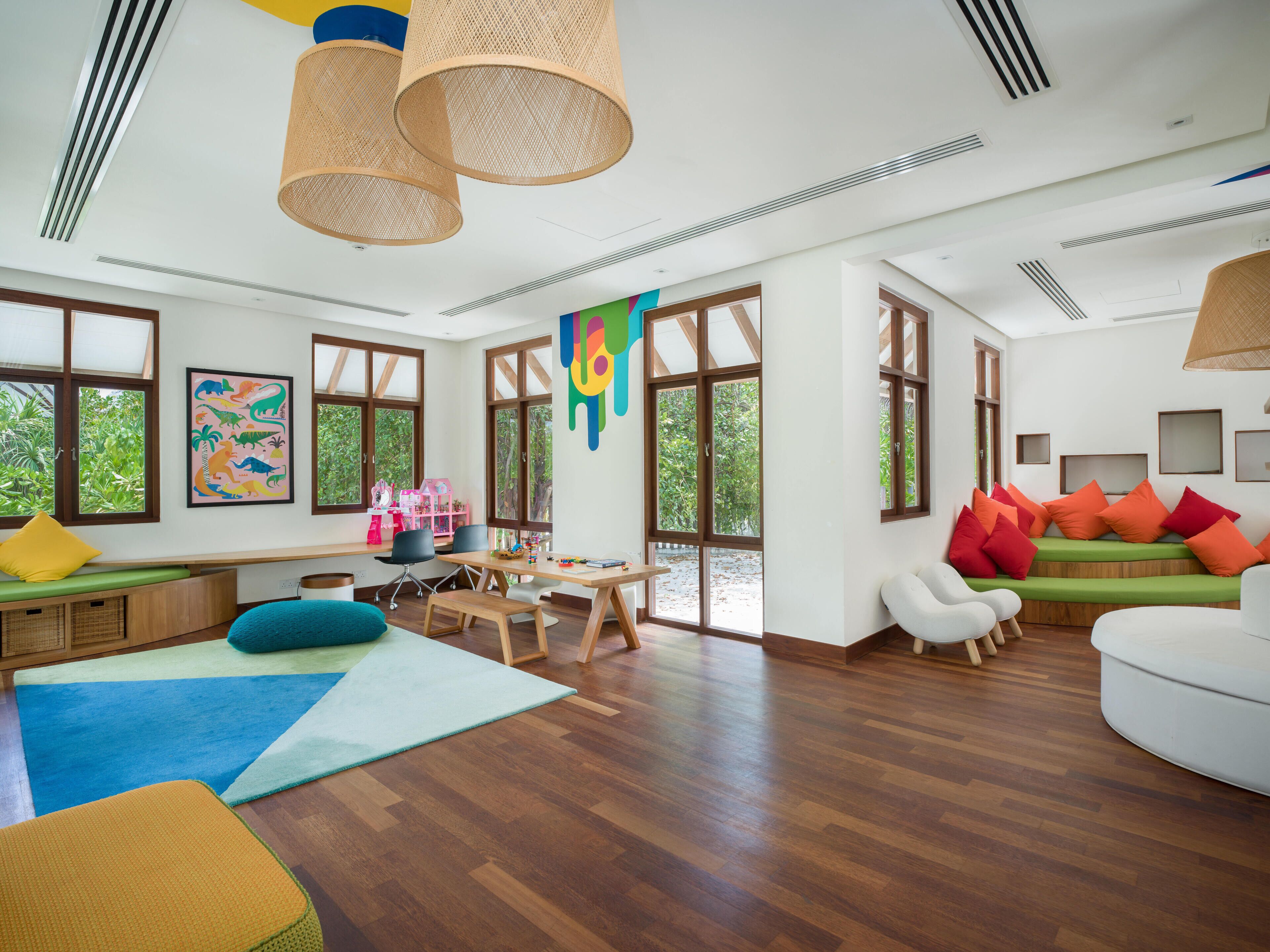 A spacious room with large windows, a wooden floor, a dining area, and a seating area with colorful cushions.