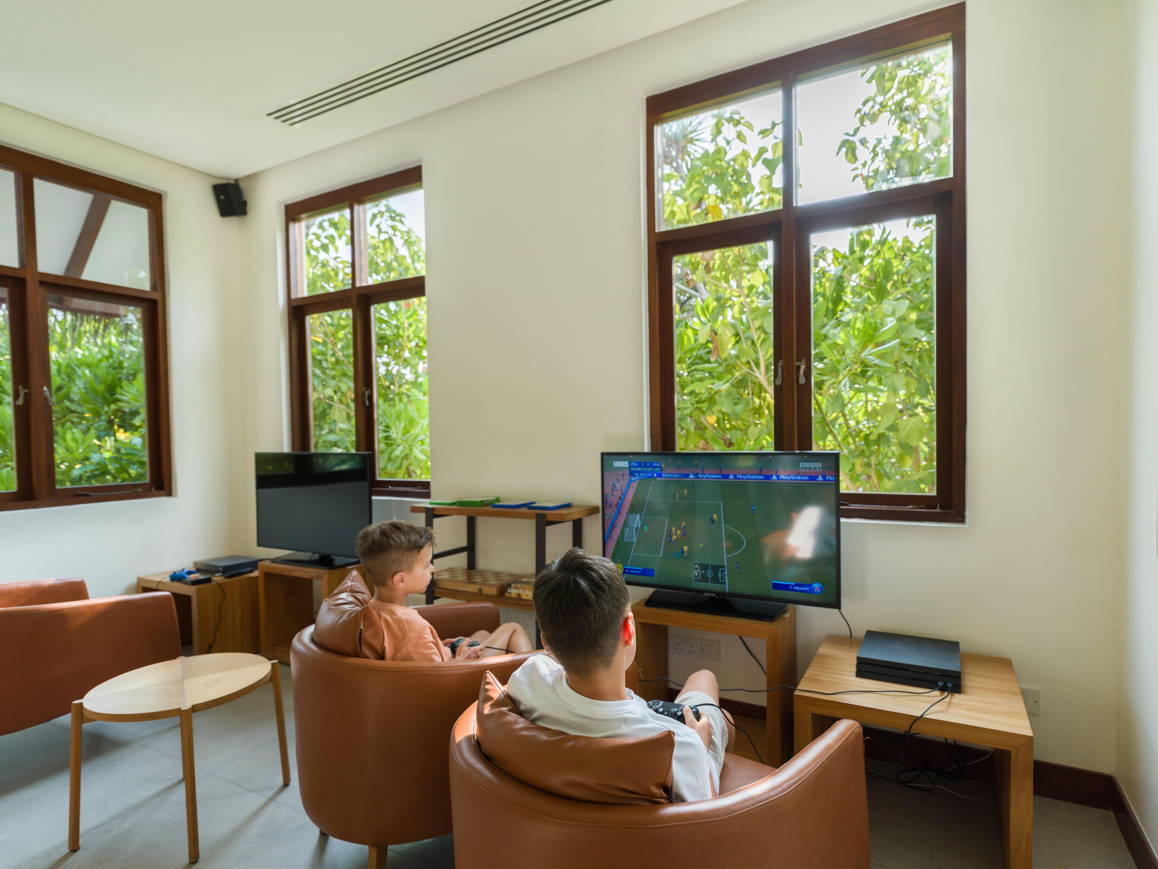 A room with two leather chairs facing a television displaying a sports game, large windows with a view of greenery, and a wooden table with a laptop.