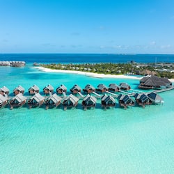Overwater bungalows offer access to the ocean via walkways, surrounded by lush greenery and a sandy beach.