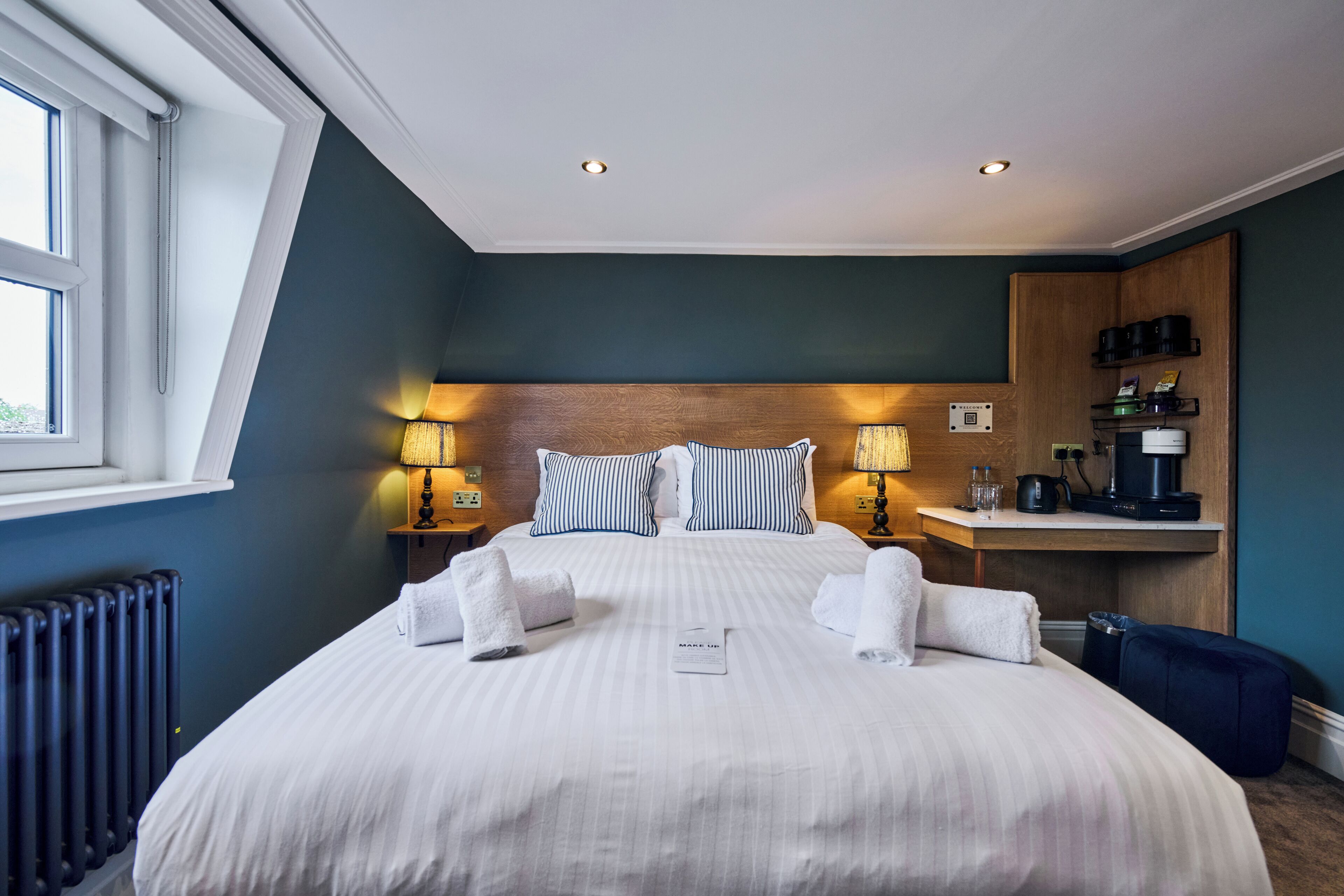 Habitación de hotel con una cama grande, dos lámparas de noche, un cabecero de madera, un escritorio pequeño con cafetera y una ventana con vistas.