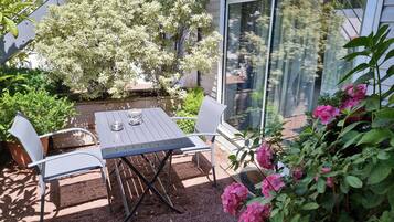 A patio with a table and chairs, surrounded by greenery and a view through a glass door.
