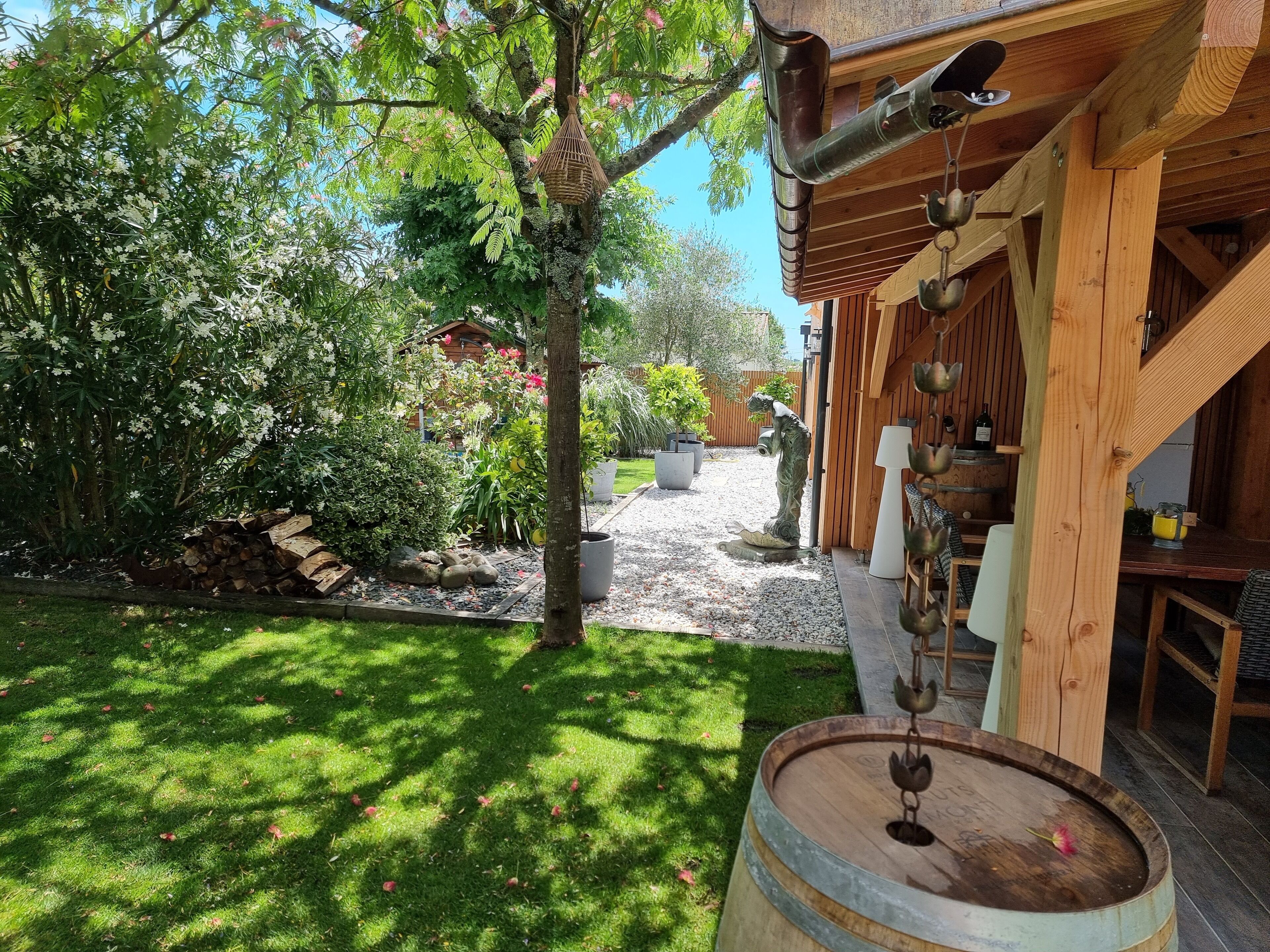 A rustic wooden building with a garden area featuring a statue, a hanging basket, and a wooden barrel.
