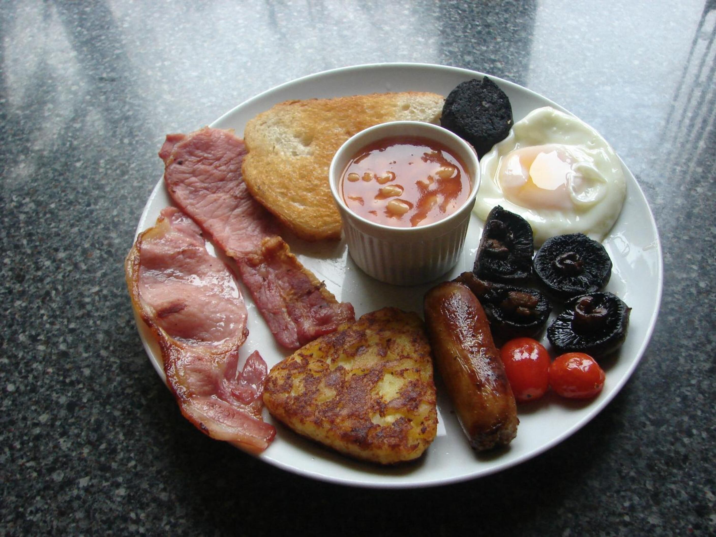 Une assiette de nourriture pour le petit-déjeuner comprenant du bacon, des saucisses, des œufs, des toasts et un petit bol de haricots.