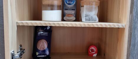 Un mueble de madera con estantes que contiene una bolsita de té, café instantáneo y un cartón de leche.