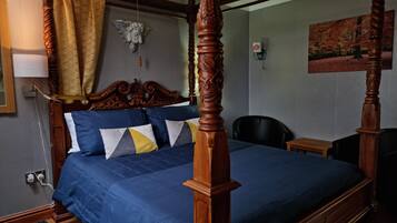 Uma cama com dossel, com colcha azul e almofadas decorativas, cabeceira de madeira, uma pequena mesa de cabeceira, uma cadeira e um quadro na parede.
