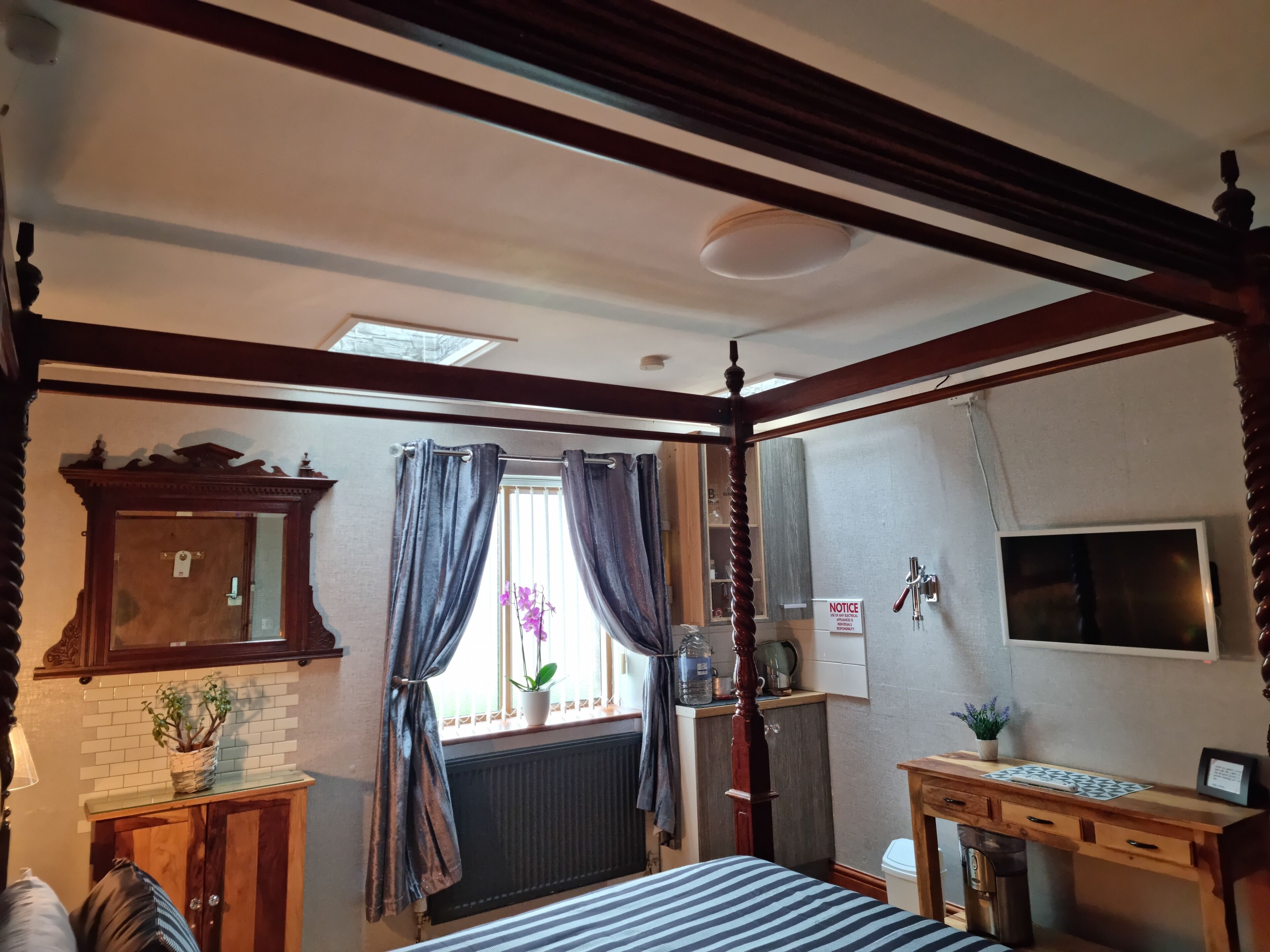 Ein Zimmer mit einem Himmelbett, einem Holzschrank, einem Heizkörper, einem Schreibtisch mit Pflanze und einem Fenster mit Vorhängen.