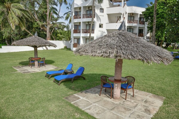 A resort with outdoor seating areas, including lounge chairs and tables, surrounded by palm trees and a building with balconies. - North Coast Beach Hotel (Mtwapa)