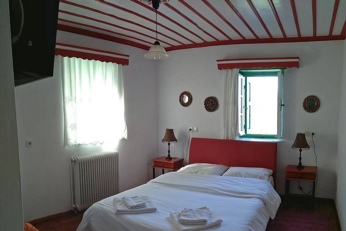 A bedroom with a bed, two bedside tables, a window with curtains, a radiator, and a ceiling fan.
