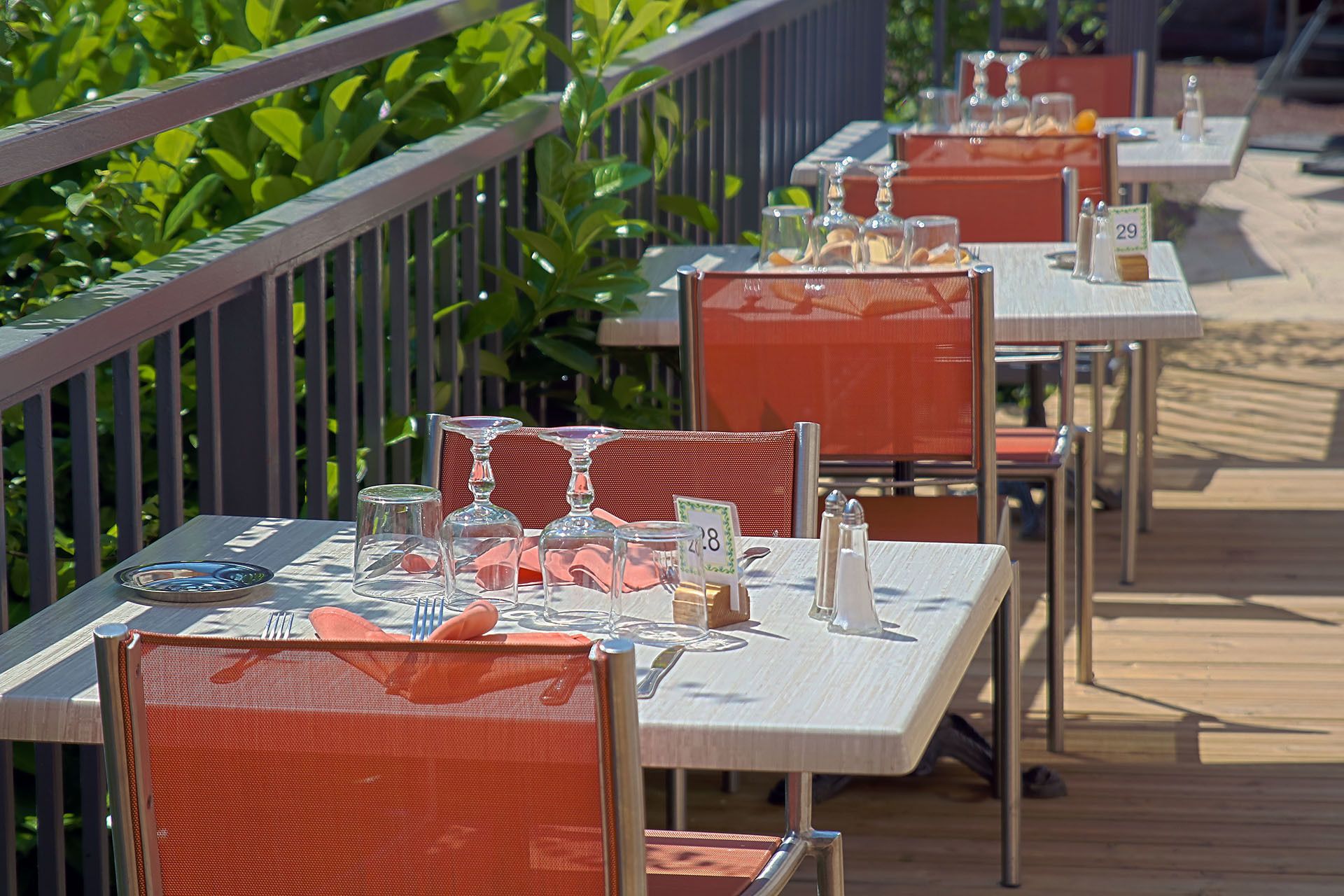 Tables et chaises pour les repas en plein air, disposées sur une terrasse en bois avec une rambarde en métal, nappées et ornées de couverts et de verres, avec une végétation luxuriante en arrière-plan.