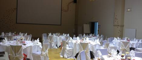 Une salle de banquet avec de nombreuses tables rondes recouvertes de nappes blanches et des chaises habillées de housses blanches.
