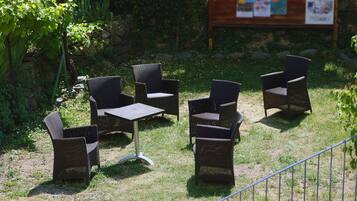 Un área de estar al aire libre con sillas de mimbre y una mesa, rodeada de follaje y un muro de piedra.