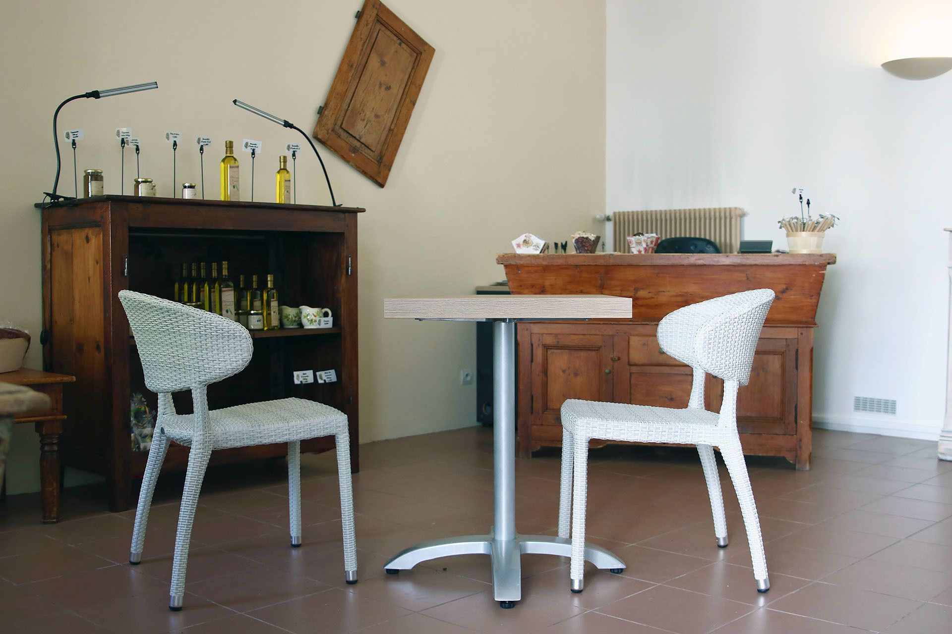 Een zithoek met twee rieten stoelen en een kleine ronde tafel. Op de muur hangt een houten kastje met flessen en een lijst met een foto.