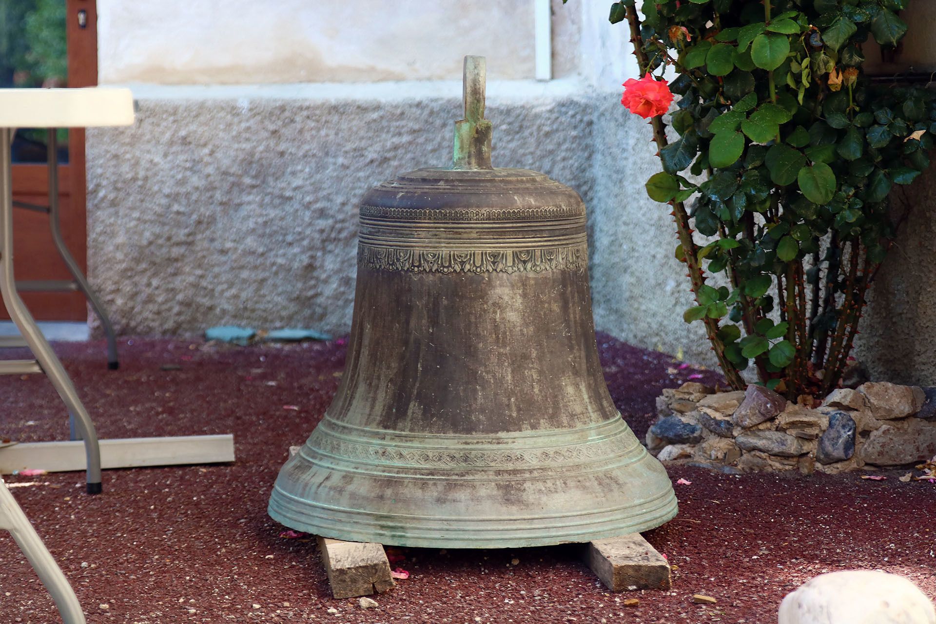 A large, weathered bell with inscriptions, placed on a wooden stand in a garden setting.