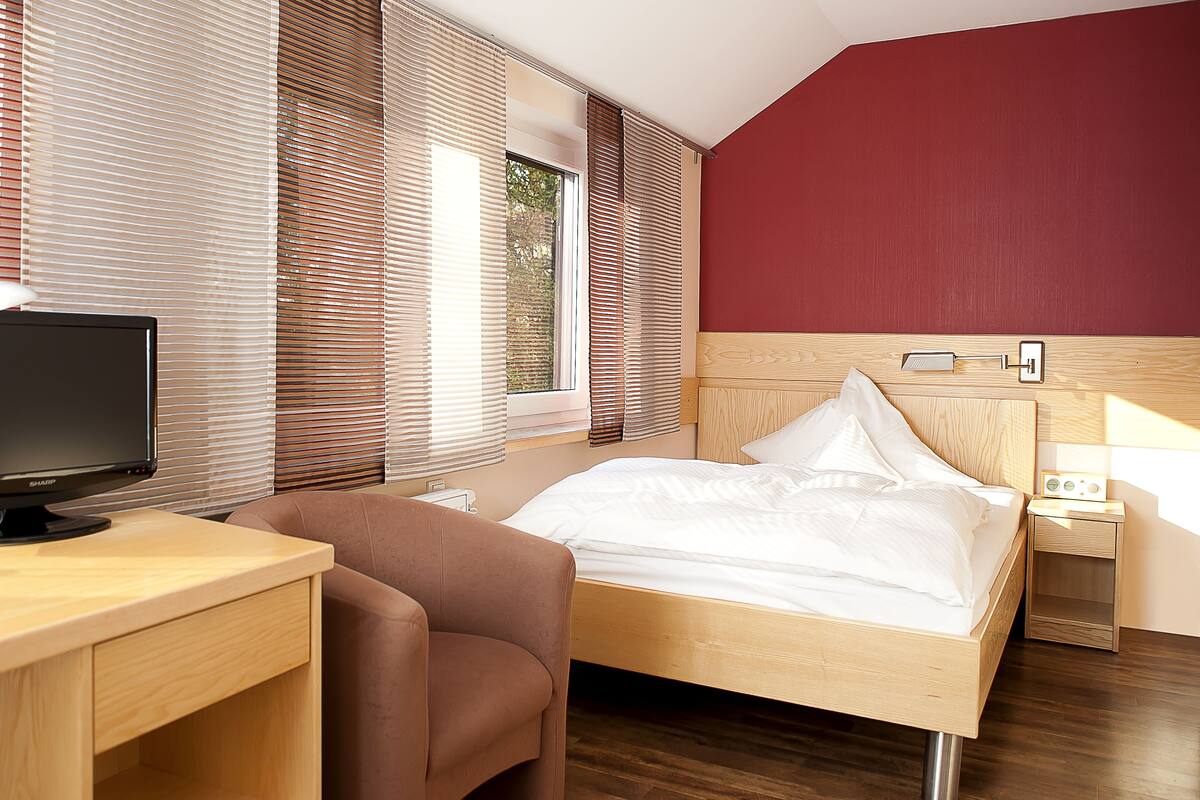 A hotel room with a wooden desk, a bed with white bedding, a brown armchair, and a nightstand with a lamp.