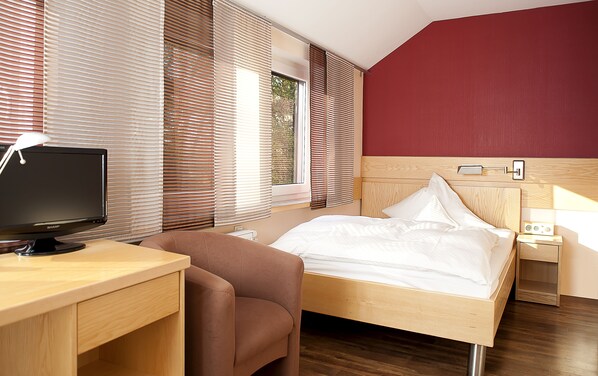 A hotel room with a wooden desk, a bed with white bedding, a brown armchair, and a nightstand with a lamp. - Pfeiffer's Sythener Flora (Haltern am See)