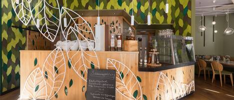 Una cafetería con mostrador de madera, un menú en una pizarra y una pared con diseño de hojas verdes.