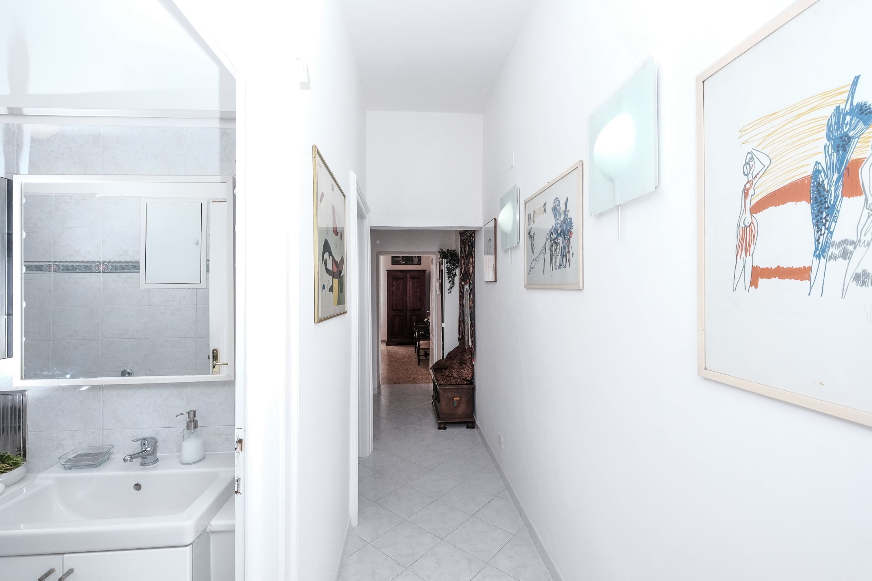 A narrow hallway with a white sink and mirror on the left, framed artwork on the walls, and a door at the end leading to another room.