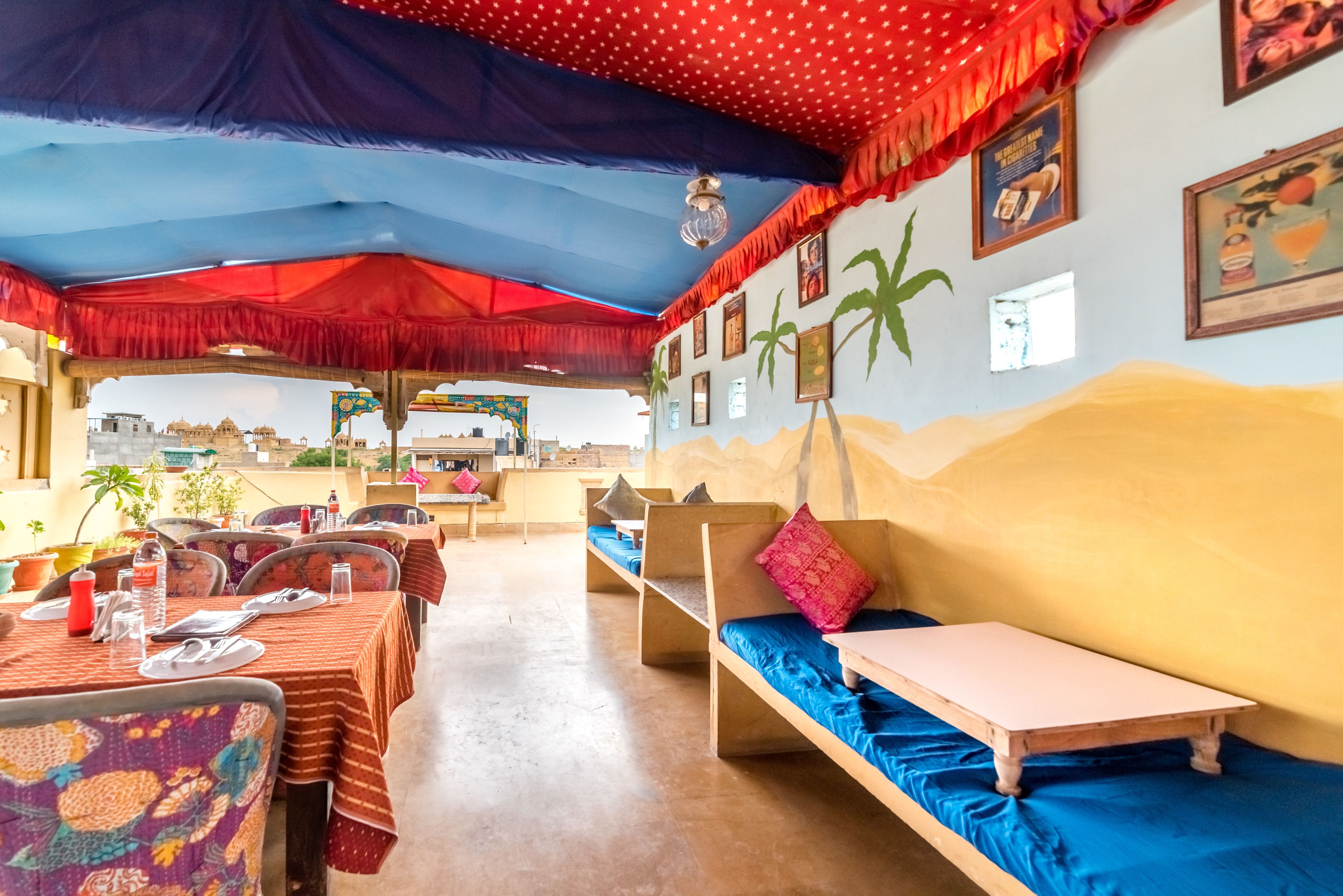 Un comedor con mesas preparadas para dos, bancos y un mural de palmeras en la pared.