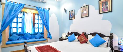 Un dormitorio con cama, cortinas azules, un asiento junto a la ventana, aire acondicionado montado en la pared y cuadros enmarcados.