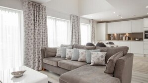 Una sala de estar moderna con un sofá seccional gris, una mesa de centro de vidrio y una cocina al fondo.